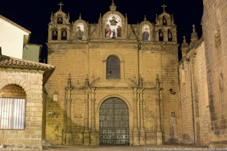 Floodlit stone church façade with large wooden door, saint niches, bell openings, and crosses.