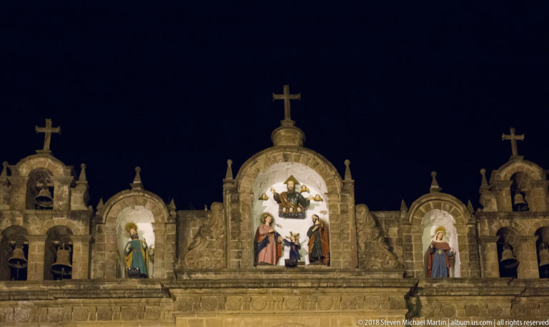 Upper church façade with three painted religious niches, crosses, bell openings, and carved stone ornament.