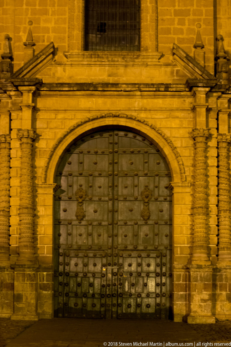 Large studded double wooden doors set within an ornate floodlit stone church portal at night.