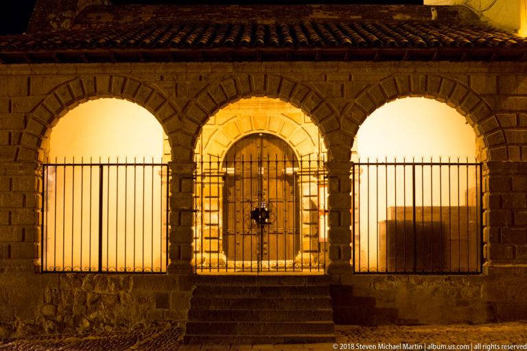 Three illuminated stone arches with iron grilles and a recessed central doorway beneath a tiled roof.
