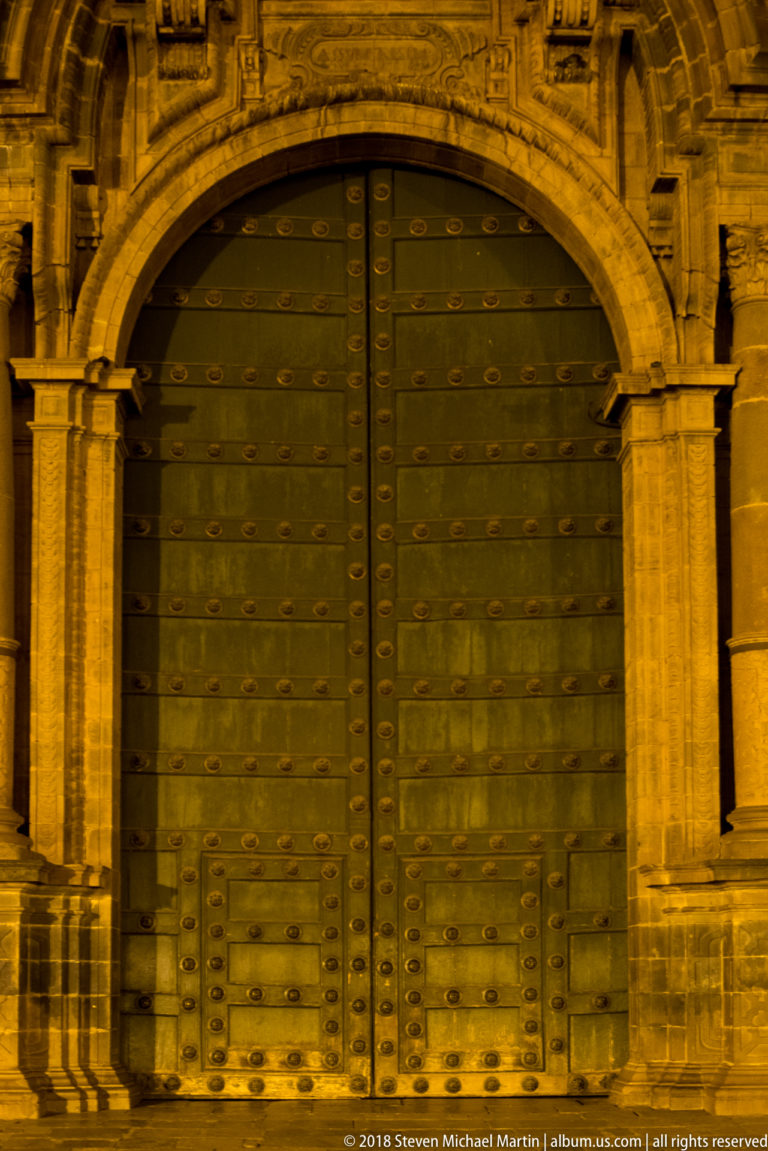 Green studded double doors beneath an ornate carved stone arch and paired columns at night.