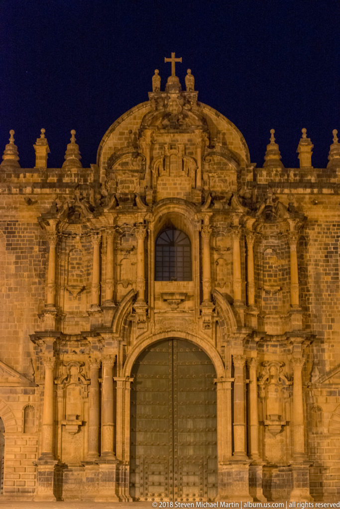 Central portal of Cusco Cathedral with massive doors, paired columns, carved stonework, and cross at night.