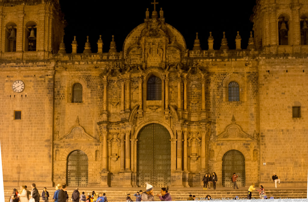 Frontal night view of Cusco Cathedral with ornate central portal, stone walls, tower, clock, and visitors below.