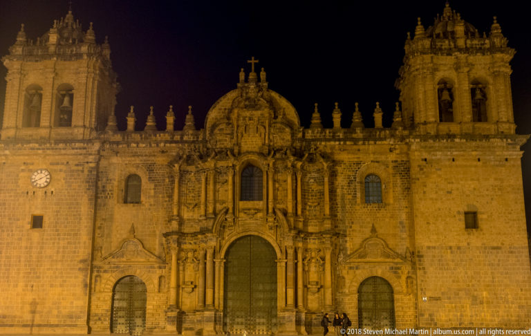 Wide illuminated view of Cusco Cathedral façade and flanking towers against a dark blue night sky.