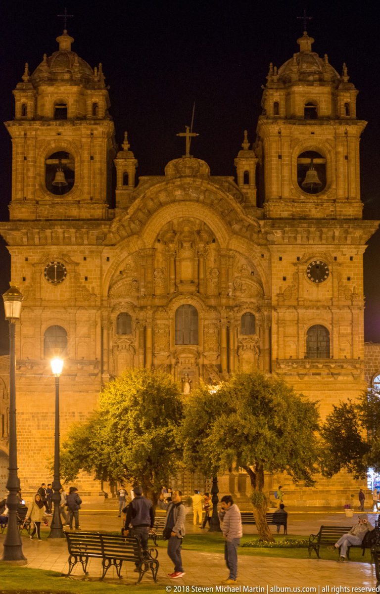 Illuminated Church of the Society of Jesus behind trees, benches, and pedestrians on Cusco’s Plaza de Armas.