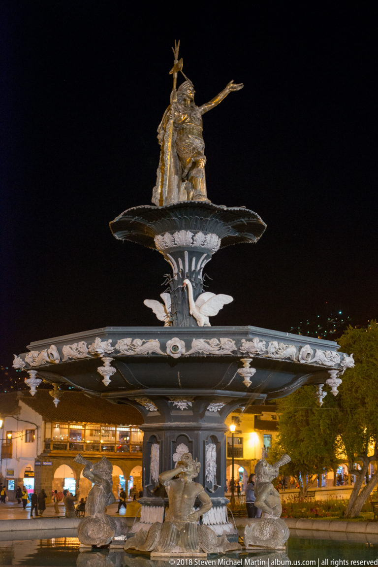 Tiered Plaza de Armas fountain topped by a gilded Pachacuti figure, illuminated against the night sky.