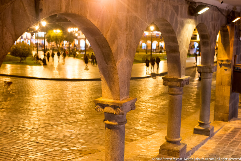 Stone arches and columns frame pedestrians crossing a wet, illuminated plaza at night in Cusco.