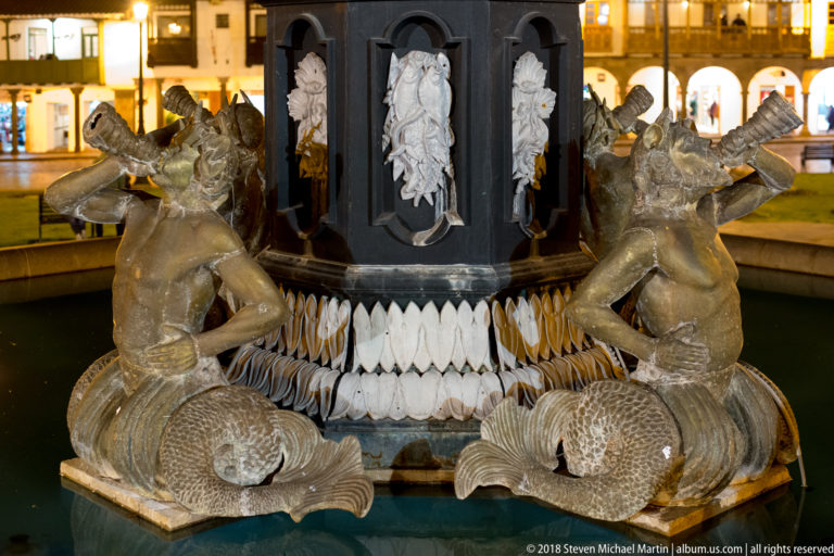 Close view of paired sculpted aquatic figures with curled scaled tails around the base of a fountain.