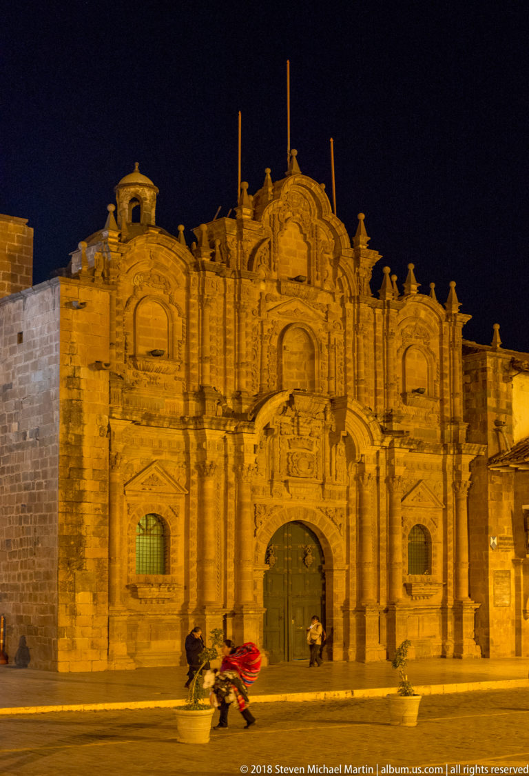 Wide floodlit colonial church façade with central doorway, flanking bays, carved stonework, and benches at night.