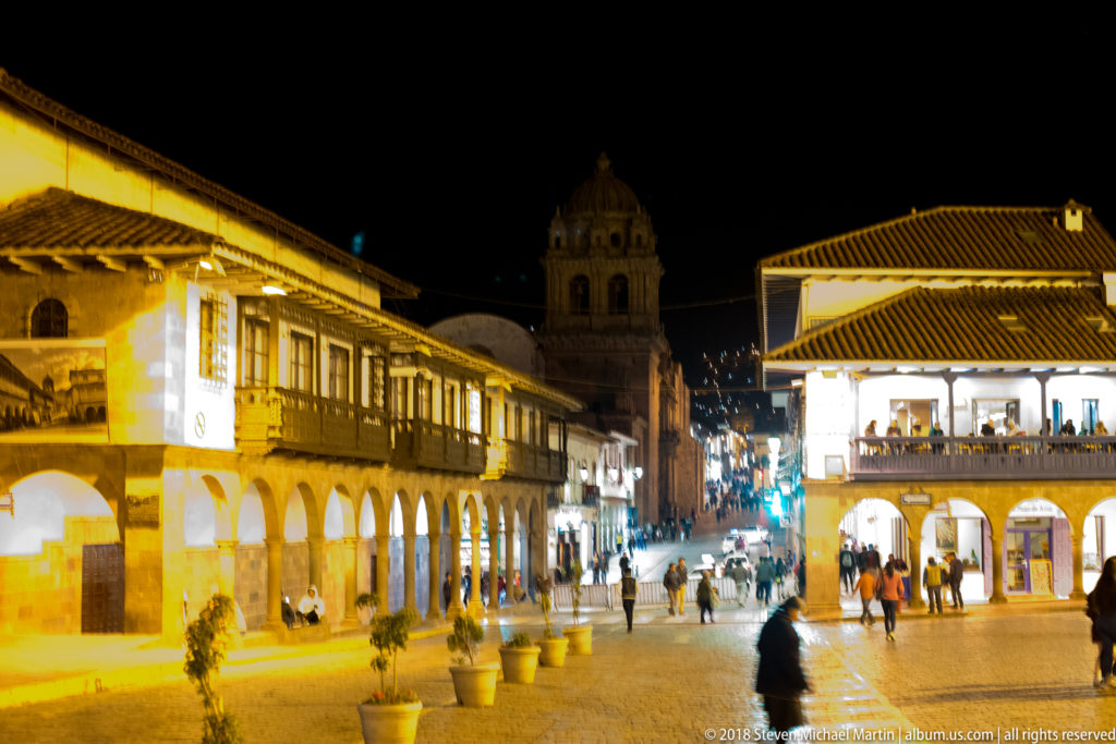 Pedestrian street between white arcaded buildings and wooden balconies, with a church tower in the distance at night.