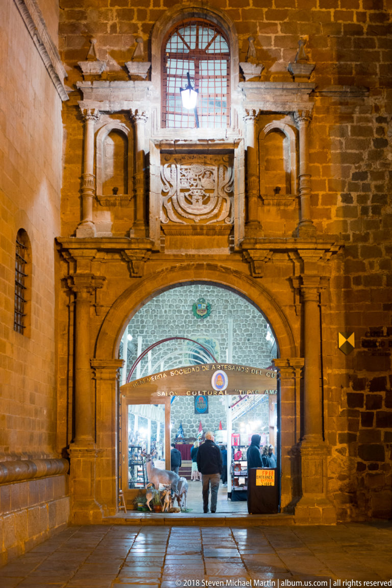 Carved stone archway in a masonry wall opening into a brightly lit indoor market with visitors.