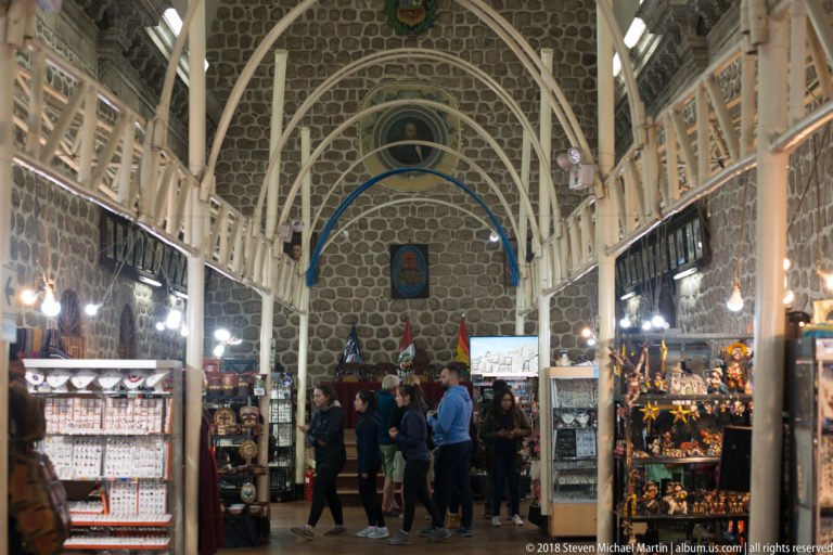 Long vaulted artisan market hall with craft stalls on both sides and visitors in the central aisle.
