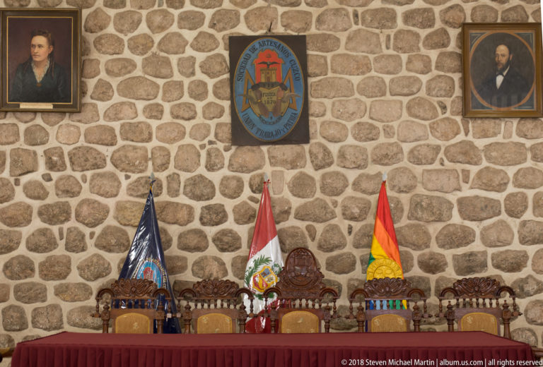 Three carved chairs behind a red table beneath institutional emblems and Peruvian and regional flags on a stone wall.