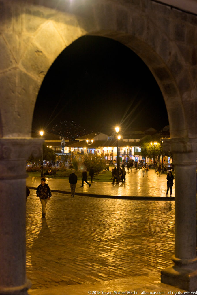 Large dark stone arch framing a wet, illuminated plaza with scattered pedestrians at night.