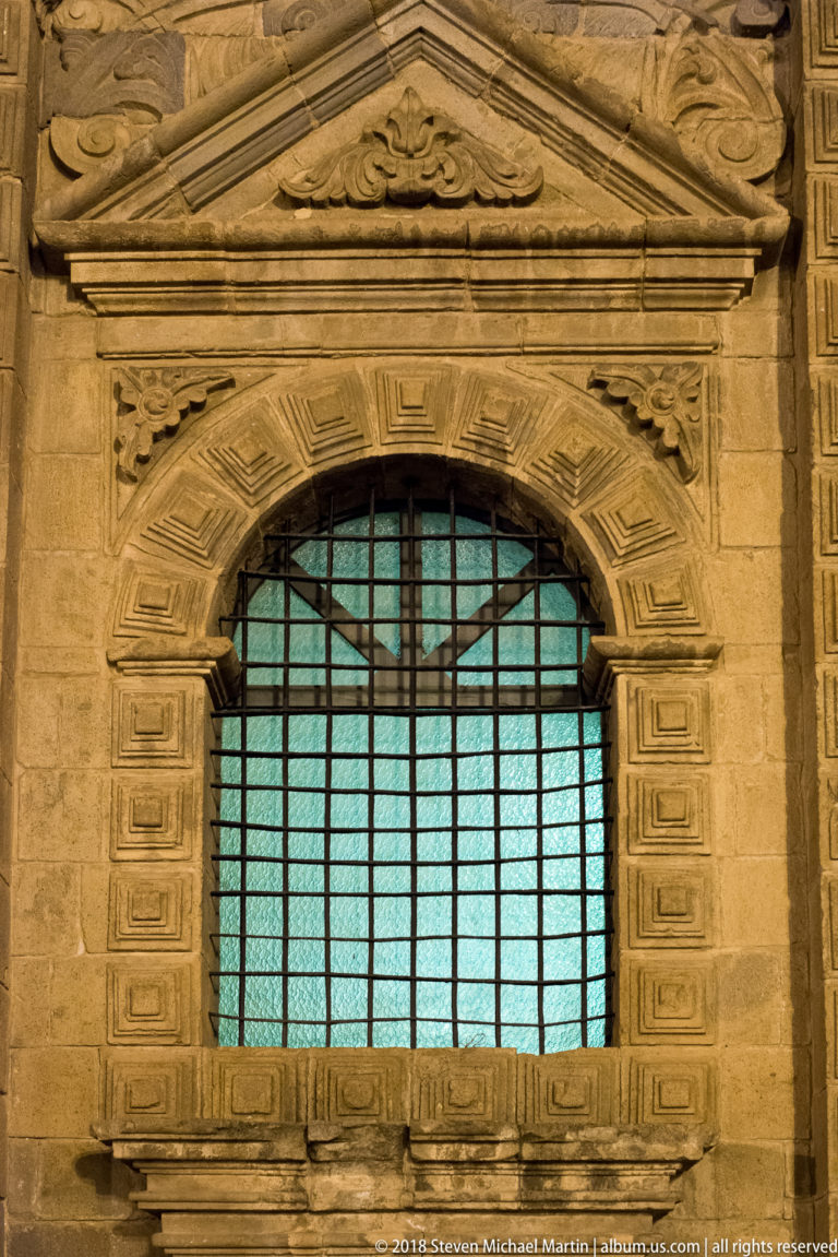 Arched barred window with turquoise panes framed by carved stone blocks and a shallow triangular pediment.