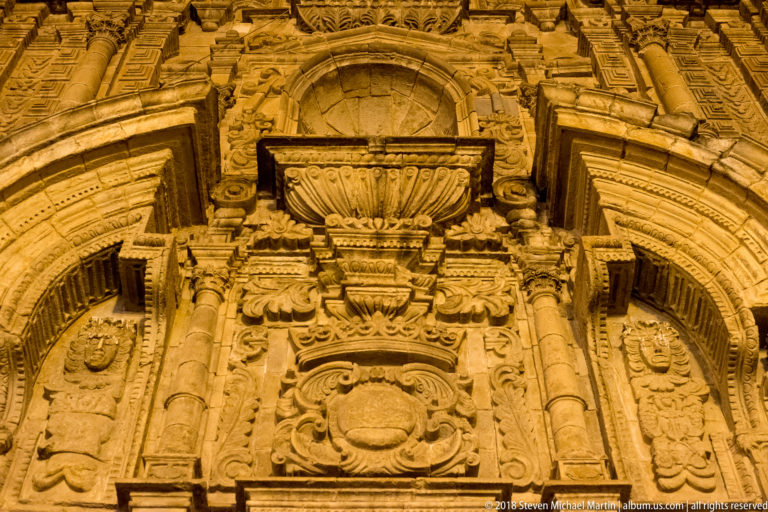 Floodlit carved stone façade with columns, scrollwork, niches, and layered Baroque ornament viewed from below.