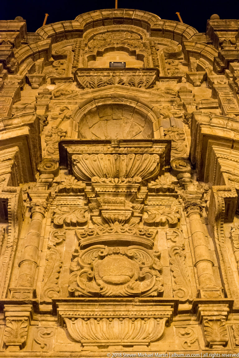 Steep upward view of a floodlit carved stone façade with stacked columns, cornices, scrolls, and reliefs.