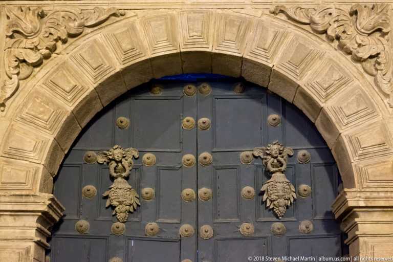 Dark arched double door with rows of metal bosses and ornate knockers in a carved stone surround.