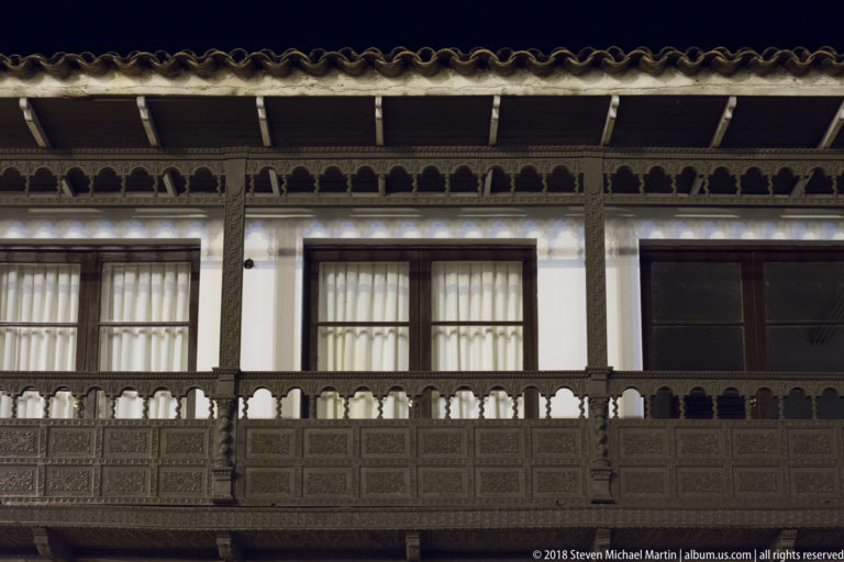 Long dark carved-wood balcony across a white façade beneath a tiled roof at night.