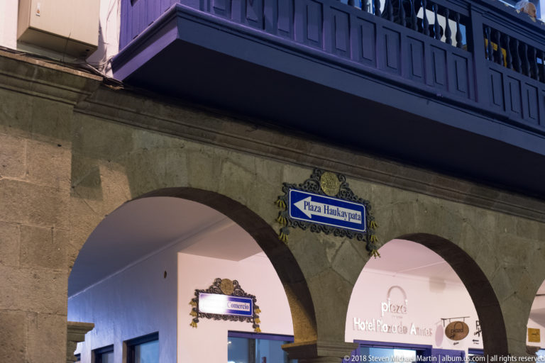 Stone arcade beneath a dark wooden balcony with a ceramic sign reading Plaza Haukaypata.