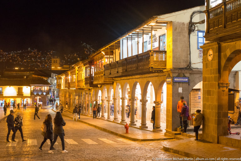 Illuminated two-story arcade with wooden balconies, pedestrians, cobbles, and hillside lights in Cusco.