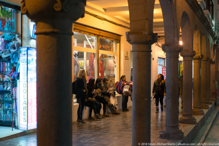 People shopping and walking beneath a stone arcade lined with brightly lit souvenir stores at night.