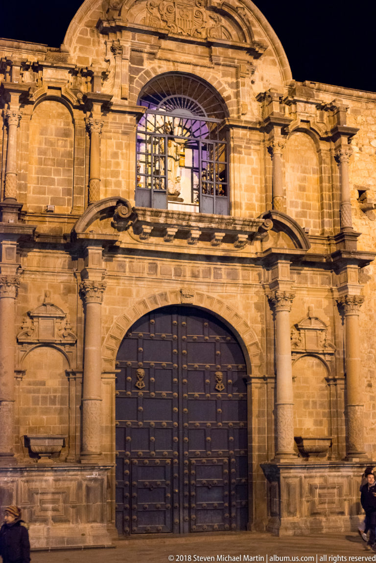 Illuminated carved-stone façade with a large studded arched door, paired columns, and an upper grilled window.