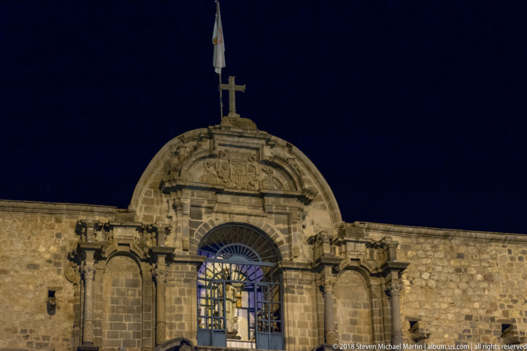 Upper floodlit stone façade with a cross, flag, tall arched grilled window, and carved architectural ornament.