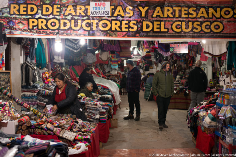 Shoppers and vendors in a narrow indoor Cusco artisan market aisle crowded with colorful textiles and souvenirs.