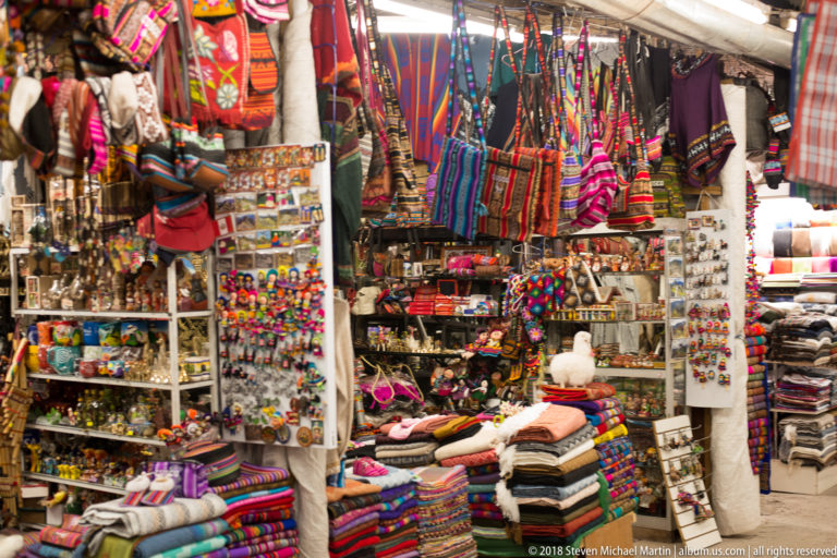 Market stall filled floor to ceiling with woven bags, folded textiles, knitwear, figurines, jewelry, and souvenirs.