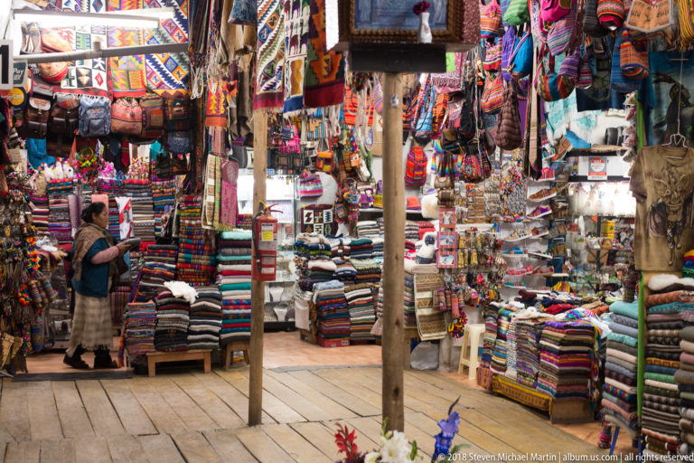 Wide view inside a Cusco artisan market with stacked textiles, hanging bags, souvenirs, wooden posts, and a shopper.
