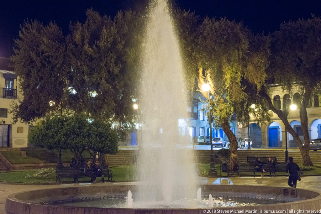 Tall illuminated fountain spraying in a tree-lined public square with benches and people at night.