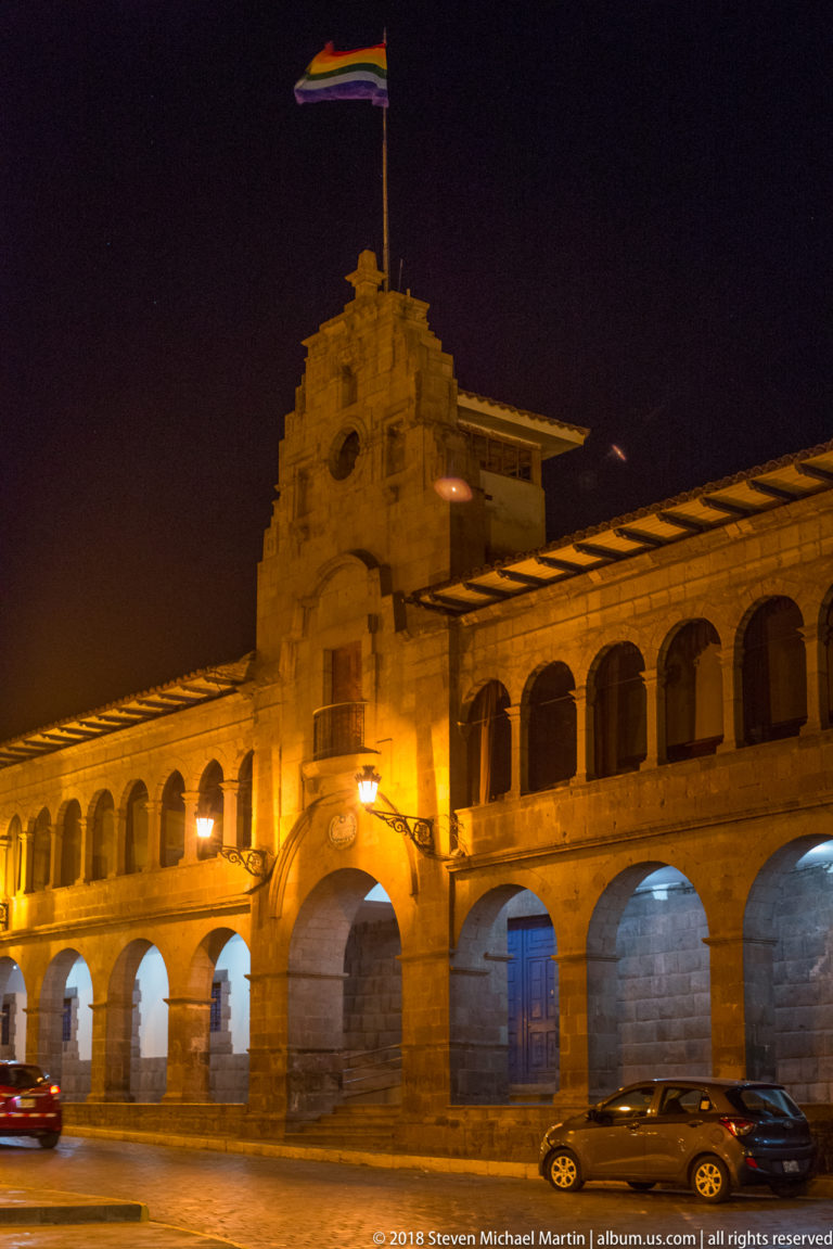 Long floodlit stone building with repeated ground-floor arches, central entrance, and Peruvian flag at night.