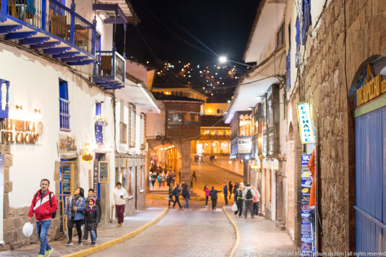 Pedestrians on a sloping Cusco street lined with white and stone façades, blue doors, shops, and hillside lights.