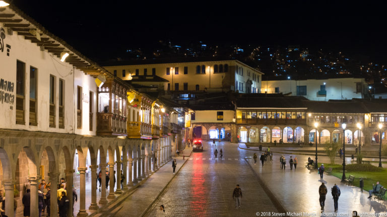 Elevated night view over an arcaded Cusco square with pedestrians and dense hillside lights beyond.