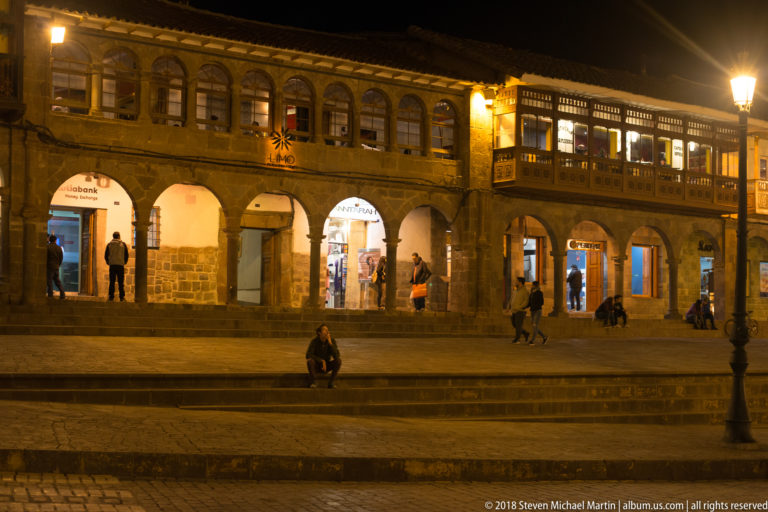 Stone commercial arcade with lit shop arches and an enclosed wooden balcony above at night.