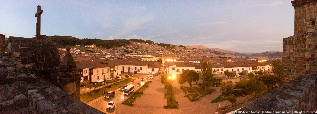 Dusk overlook from stone walls across a Cusco plaza toward densely built hills and glowing streetlights.