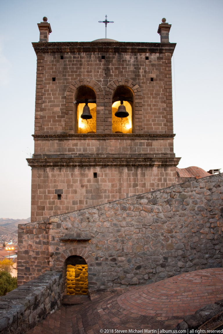 Stone bell tower with two warmly illuminated bells in paired arches above a rough masonry wall at dusk.