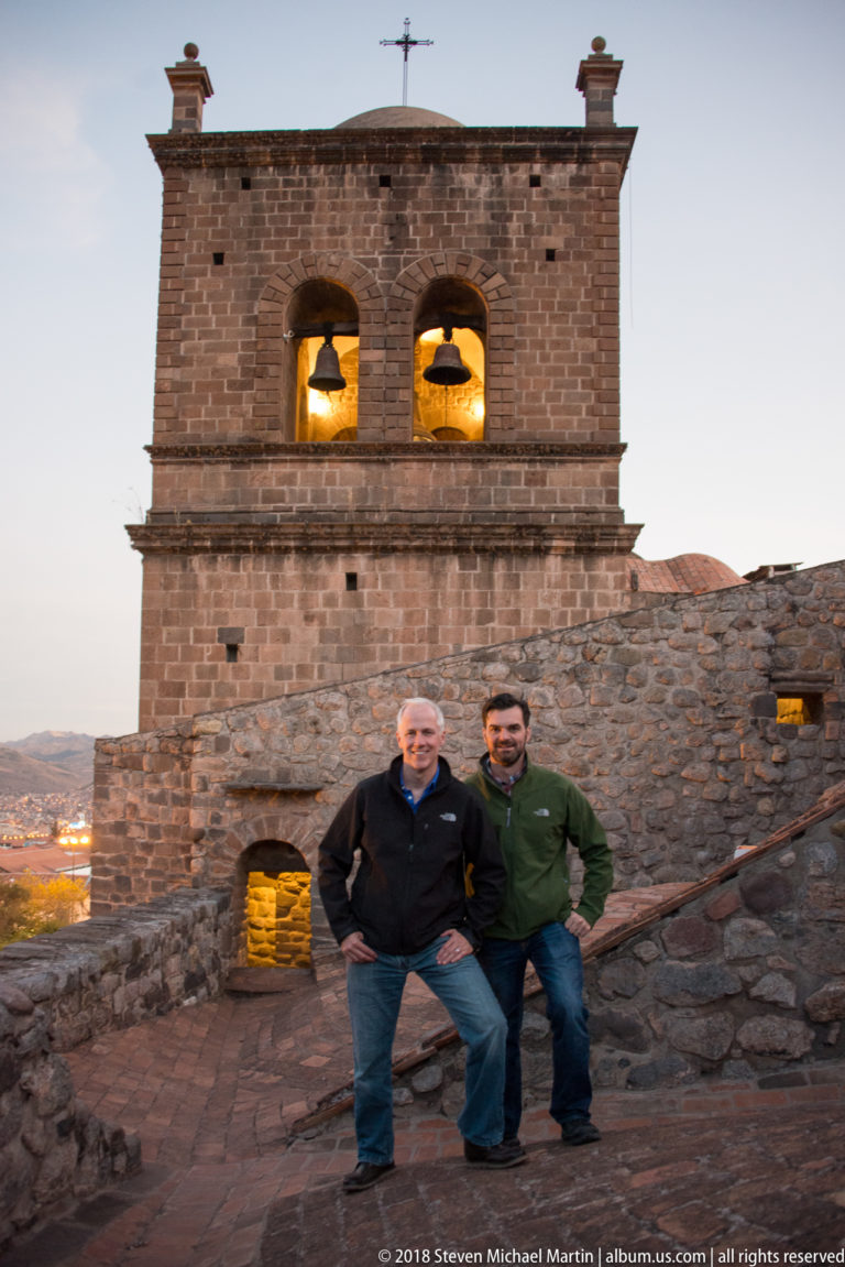 Two men posing on a stone terrace before a bell tower with two illuminated bells at dusk.