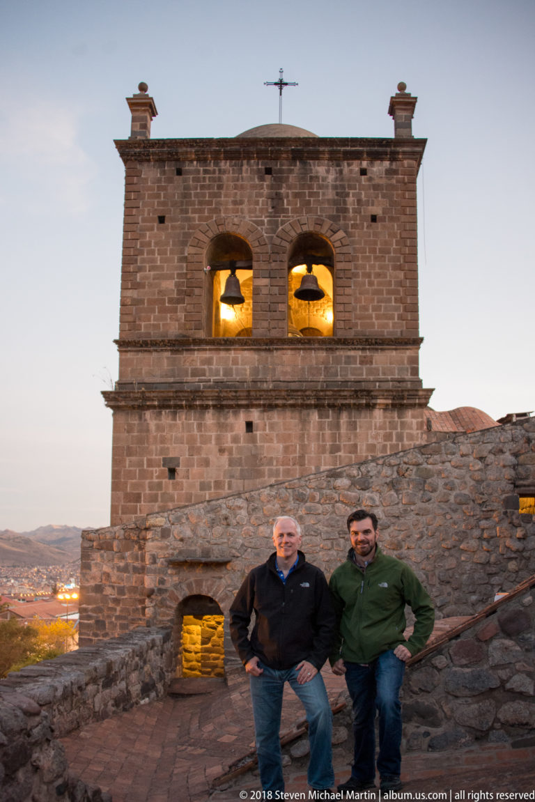 Two men standing below a stone bell tower with twin glowing bells on a sloped terrace at dusk.
