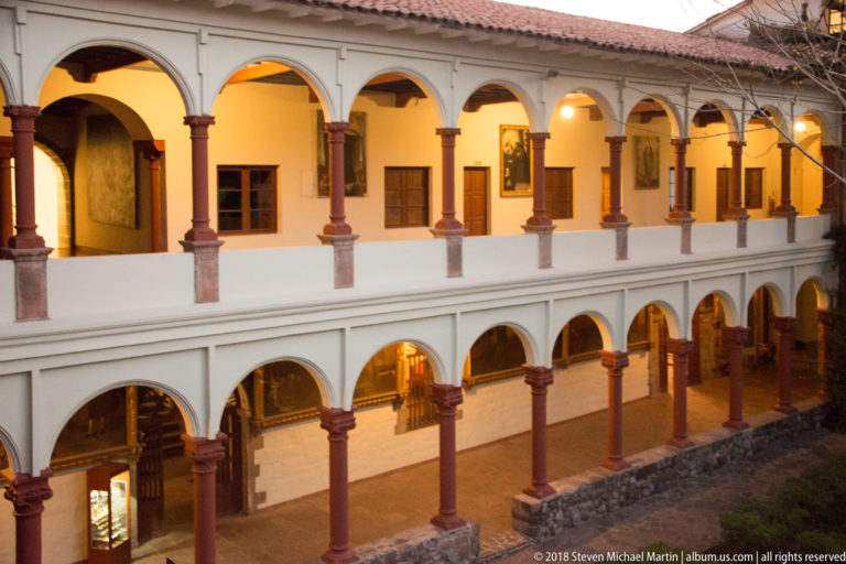 Two-story white cloister with rows of rounded arches, reddish columns, and warmly lit galleries around a courtyard.