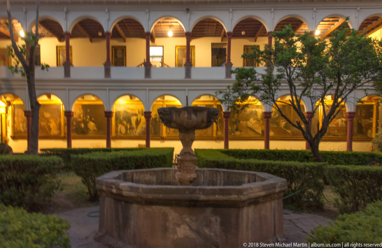 Stone fountain amid clipped hedges in a two-story arcaded courtyard glowing with warm interior light.