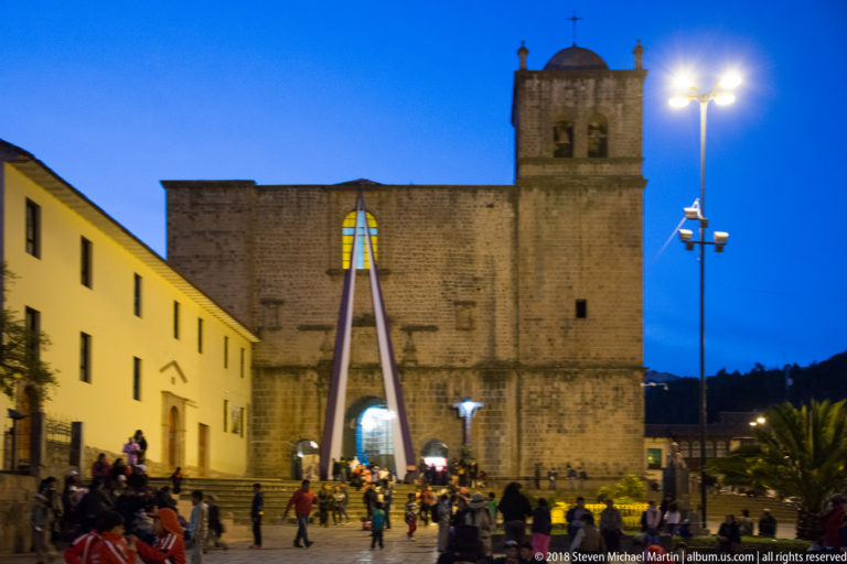 Large stone church façade facing a plaza with pedestrians and bright modern light poles under a blue evening sky.