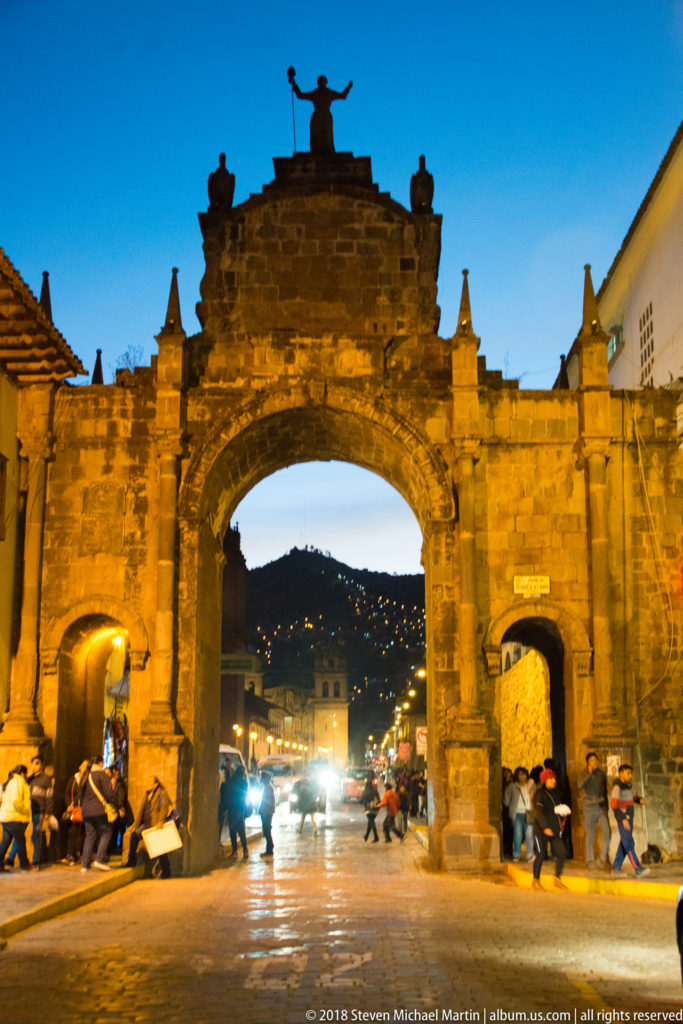 Illuminated Arco de Santa Clara spanning a busy Cusco street with pedestrians and headlights at blue hour.