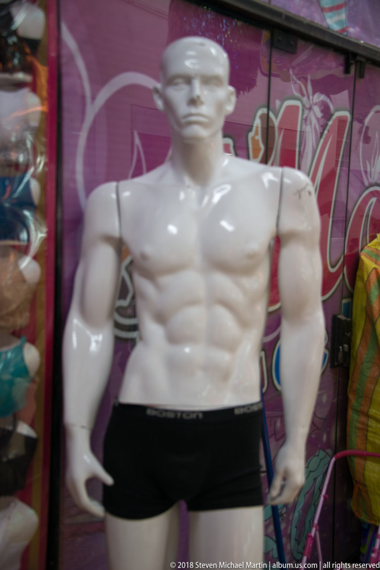 Muscular white male mannequin wearing black briefs in front of a colorful clothing-store display.