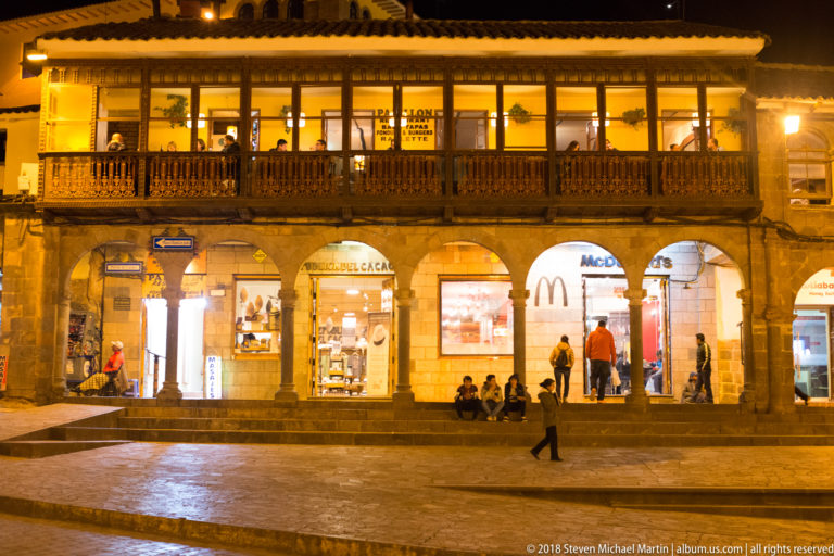 Long wooden balcony above illuminated stone arches, shops, and people along a Cusco plaza at night.