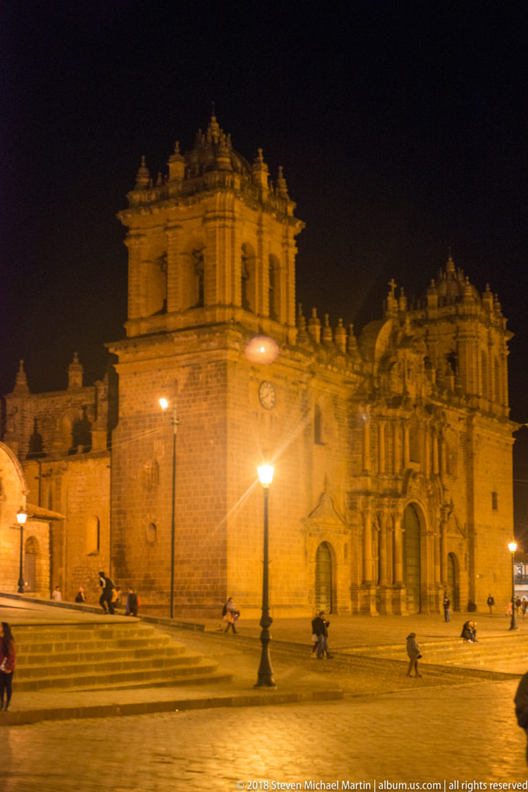Floodlit stone façade and tower of Cusco Cathedral above broad plaza steps at night.