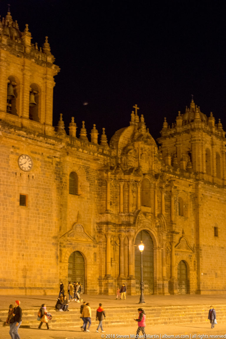 Cusco Cathedral façade illuminated at night, with clock, ornate portal, tower, and people on the steps.