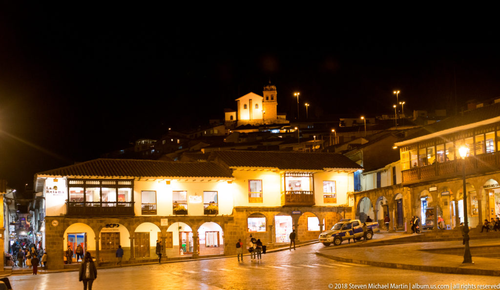 Lit arcaded buildings on a wet plaza with a small illuminated church on the hillside above.