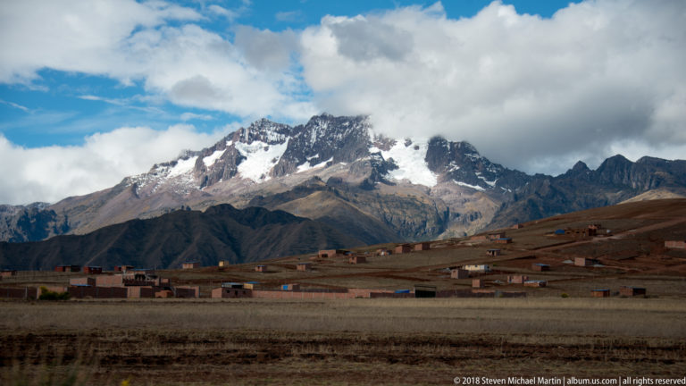 Scattered highland houses on brown fields beneath a steep, snow-streaked Andean mountain and low clouds.